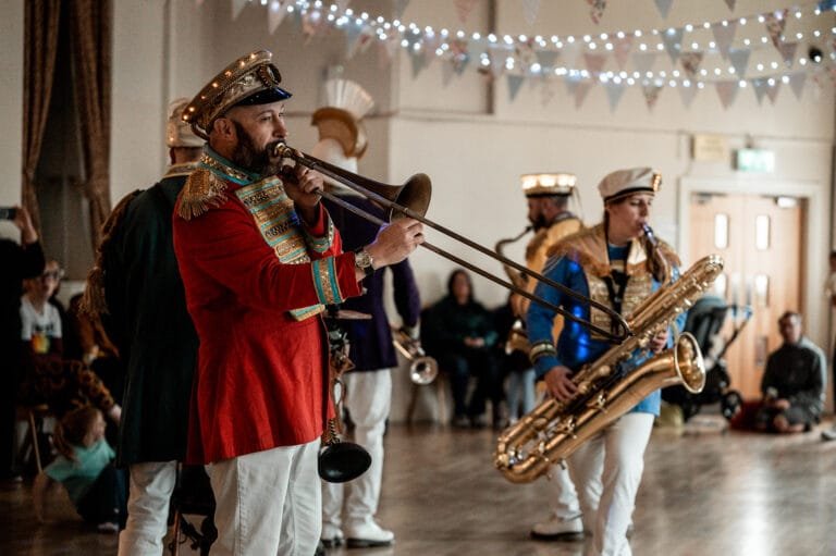 Brass band performing in a decorated indoor venue, featuring a trombone player in a vibrant red uniform, captured by Manchester event photographer Blake Hillam.